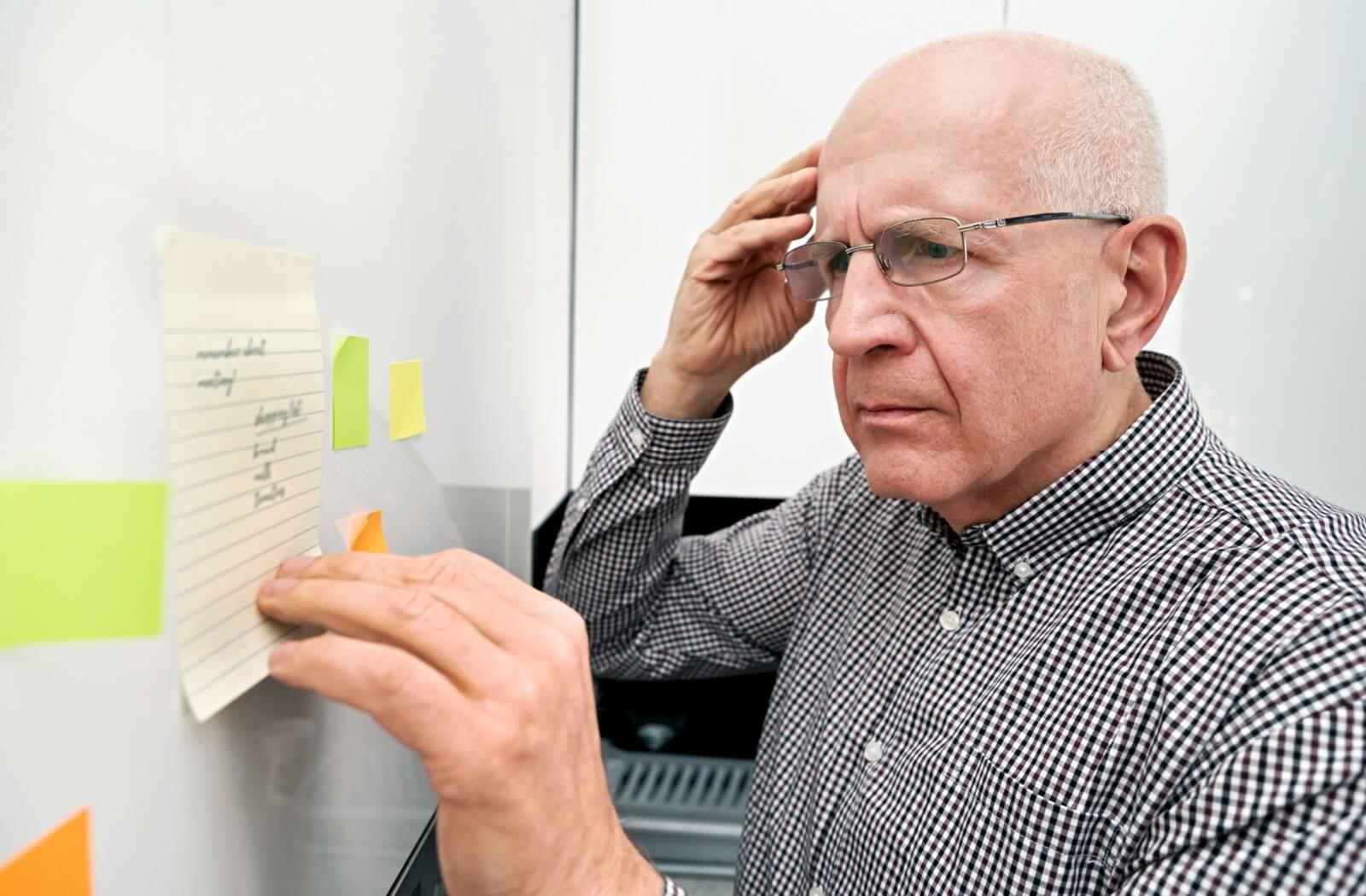 An older adult man with a confused look looking at a note posted on the kitchen cabinet.