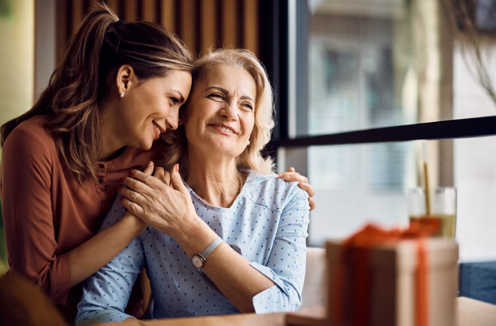 An adult daughter hugging her senior mother from behind while they both smile