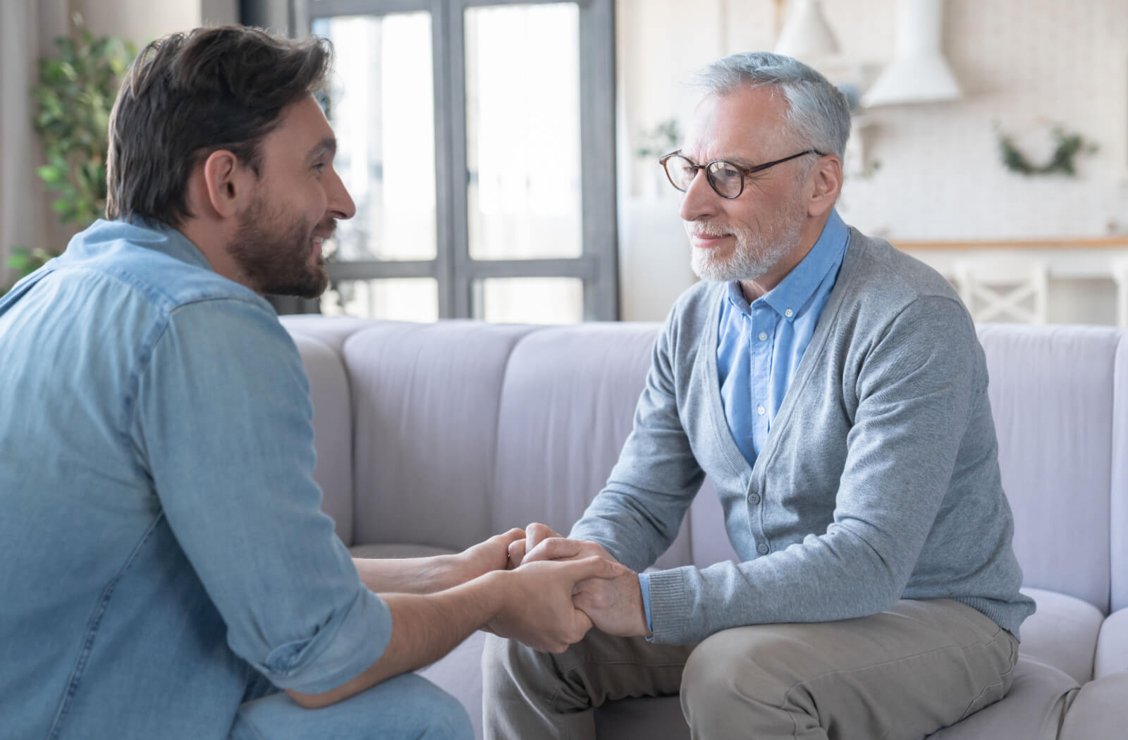 An older adult clasping hands with their adult child while sitting across from one another discussing if it's time for memory care.