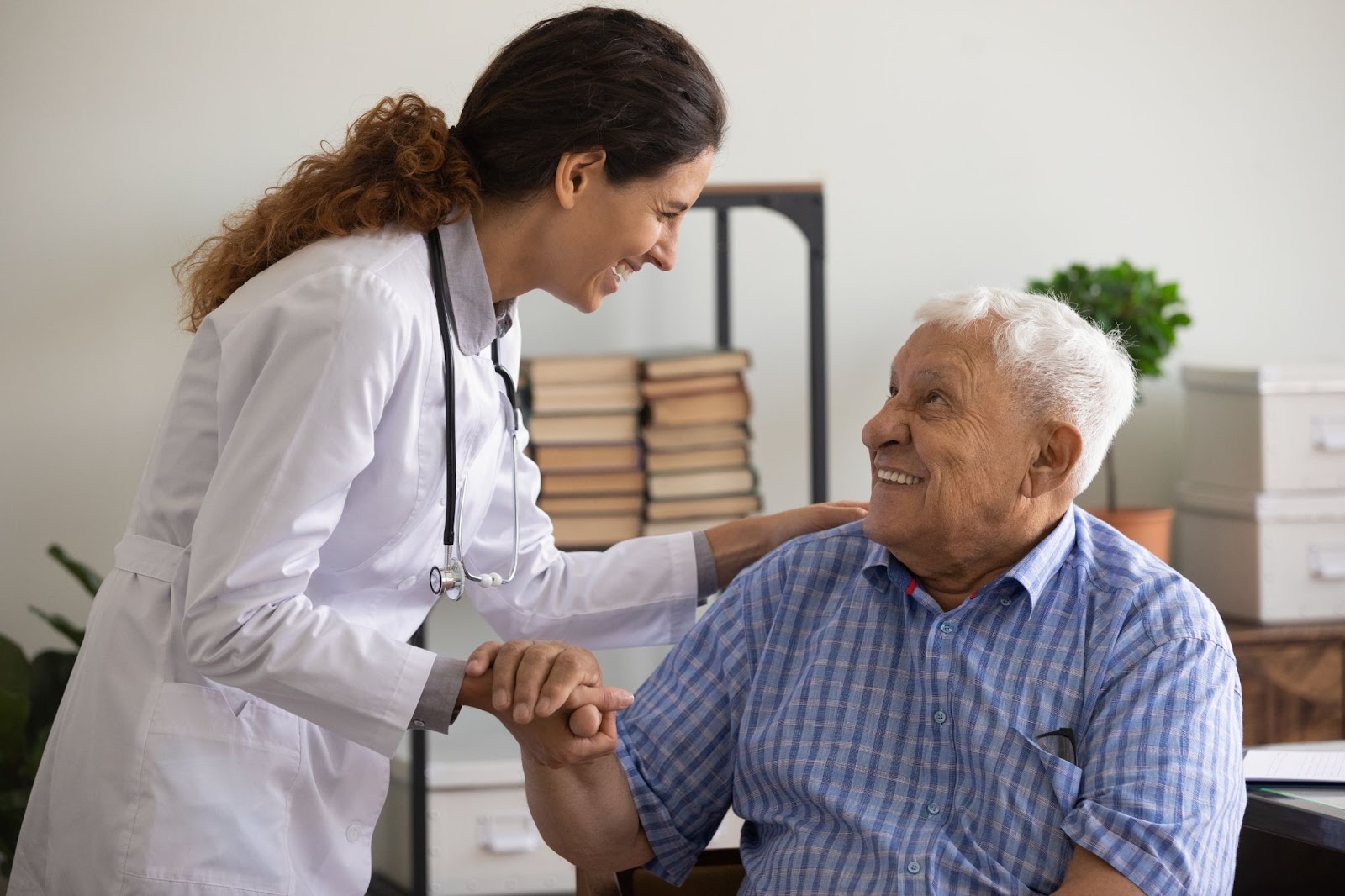 A young doctor smiling at an older male patient as he looks up at her and holds onto her hand