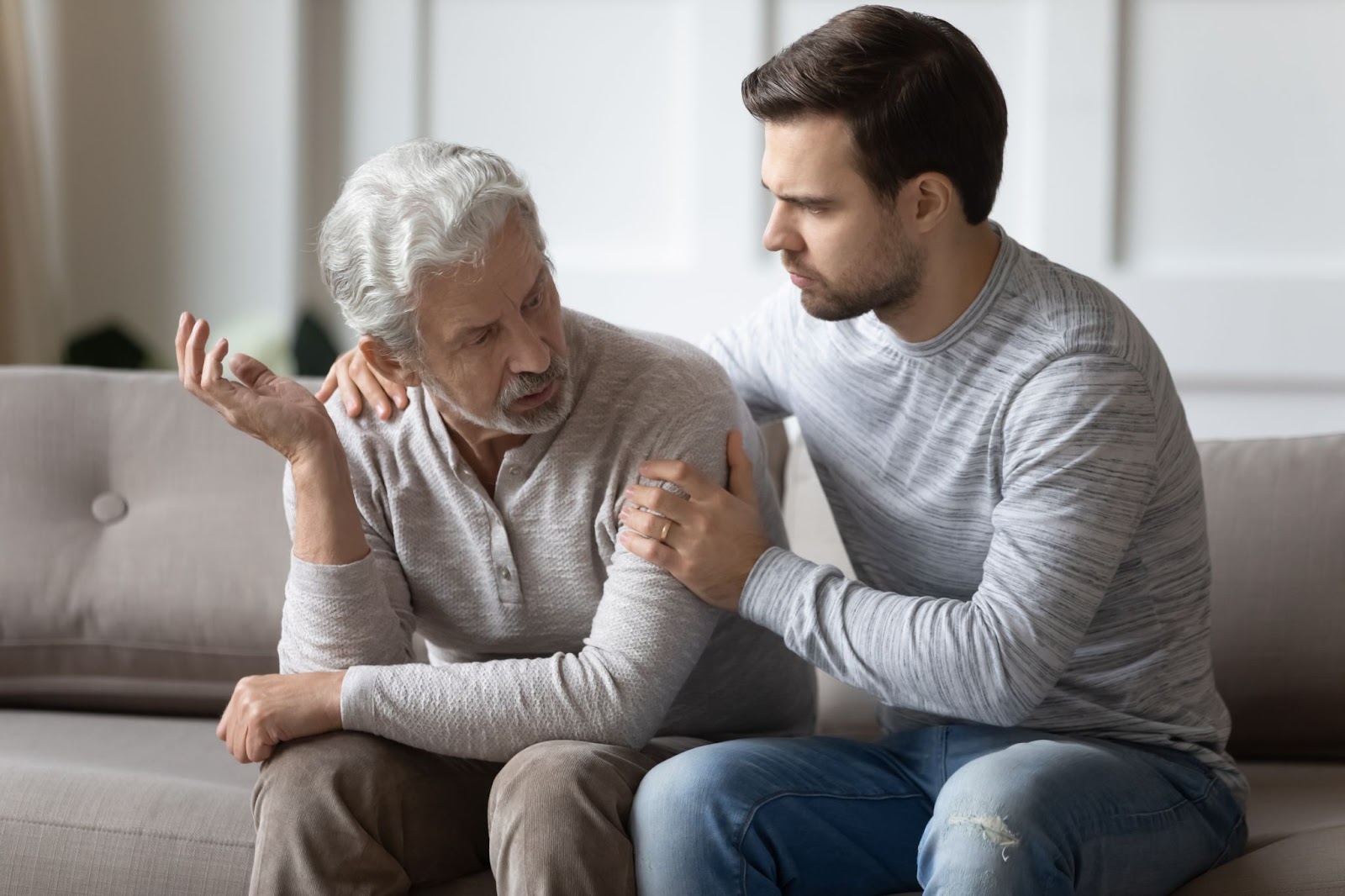 A young man sitting on the couch with his aging father and having a serious conversation.