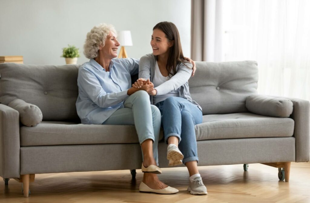 A senior mother and her adult daughter on a couch smiling and holding hands