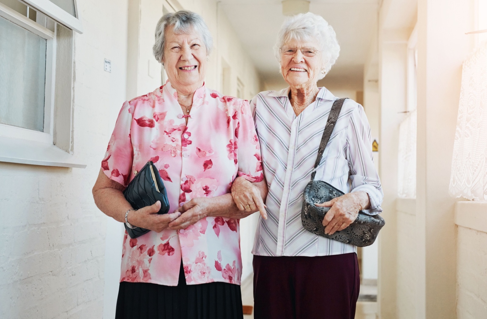 Two smiling older adults with handbags standing arm in arm.