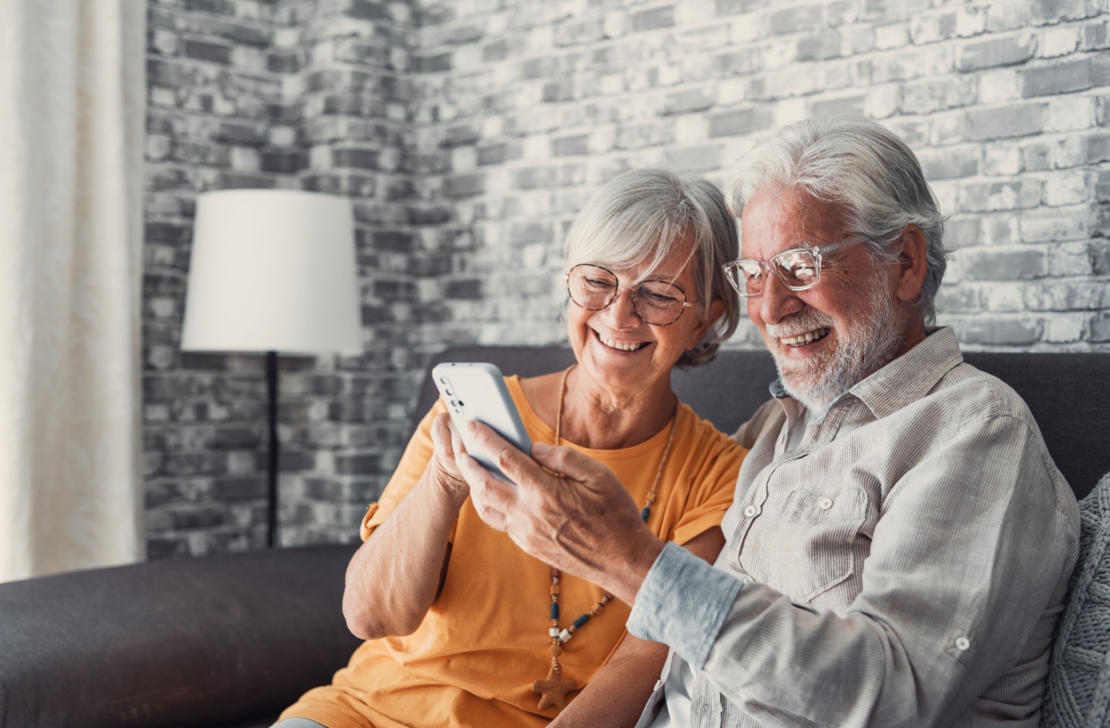 An older couple sitting on the couch in senior living, laughing and looking at their phone together.