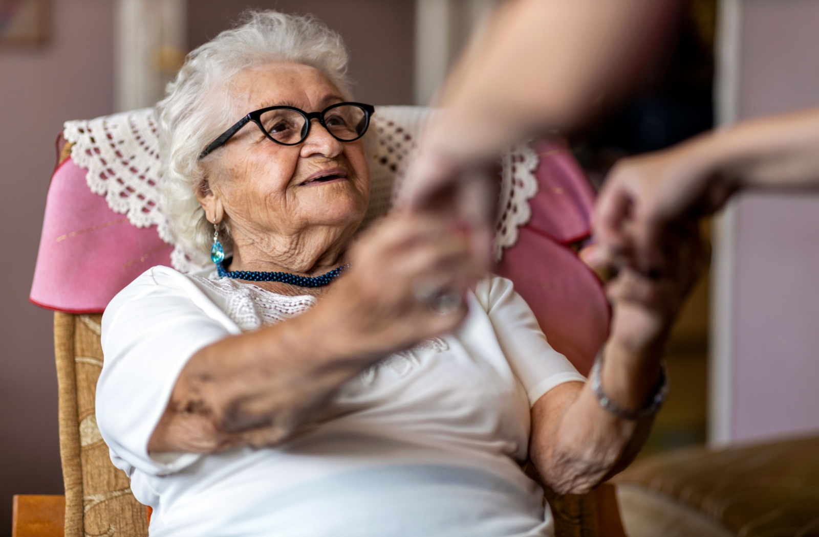 An older adult with dementia smiling as their adult child gets ready to help them from a rocking chair, holding hands.