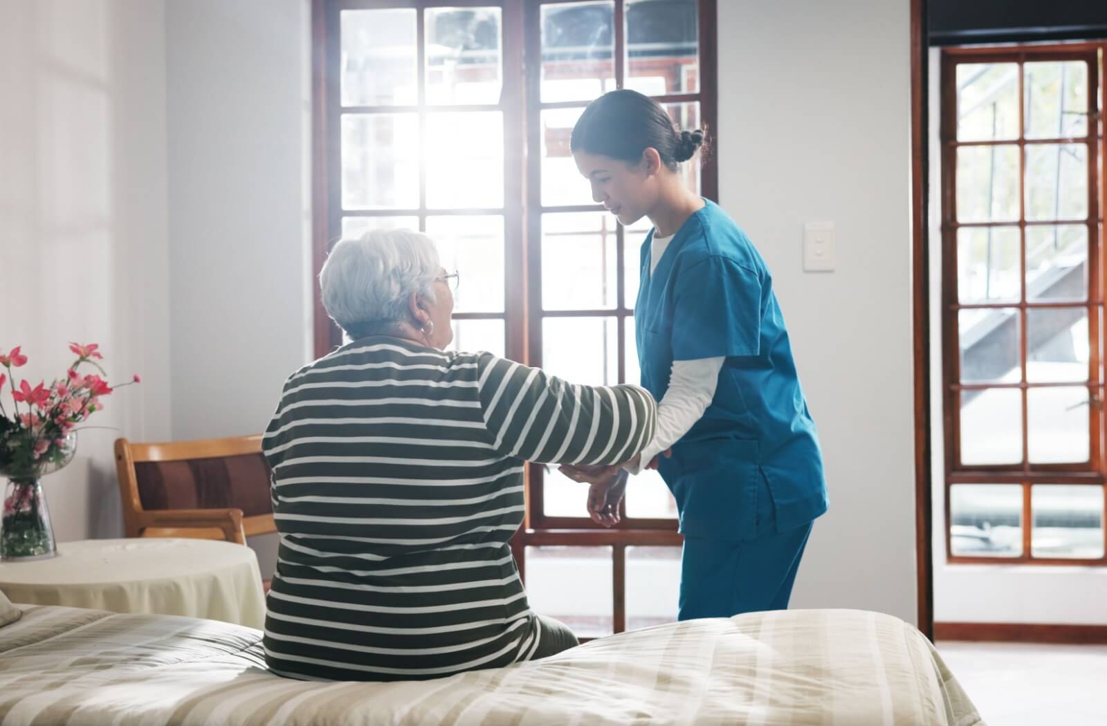 Care team member assisting older adult from bed in a bright room with natural light and supportive one-on-one attention
