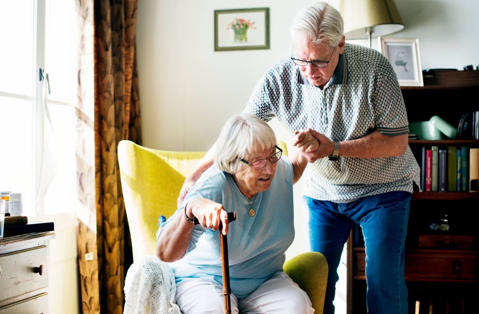 An older adult uses their cane to stand up from a yellow armchair while their spouse offers support in a living room of a senior living home