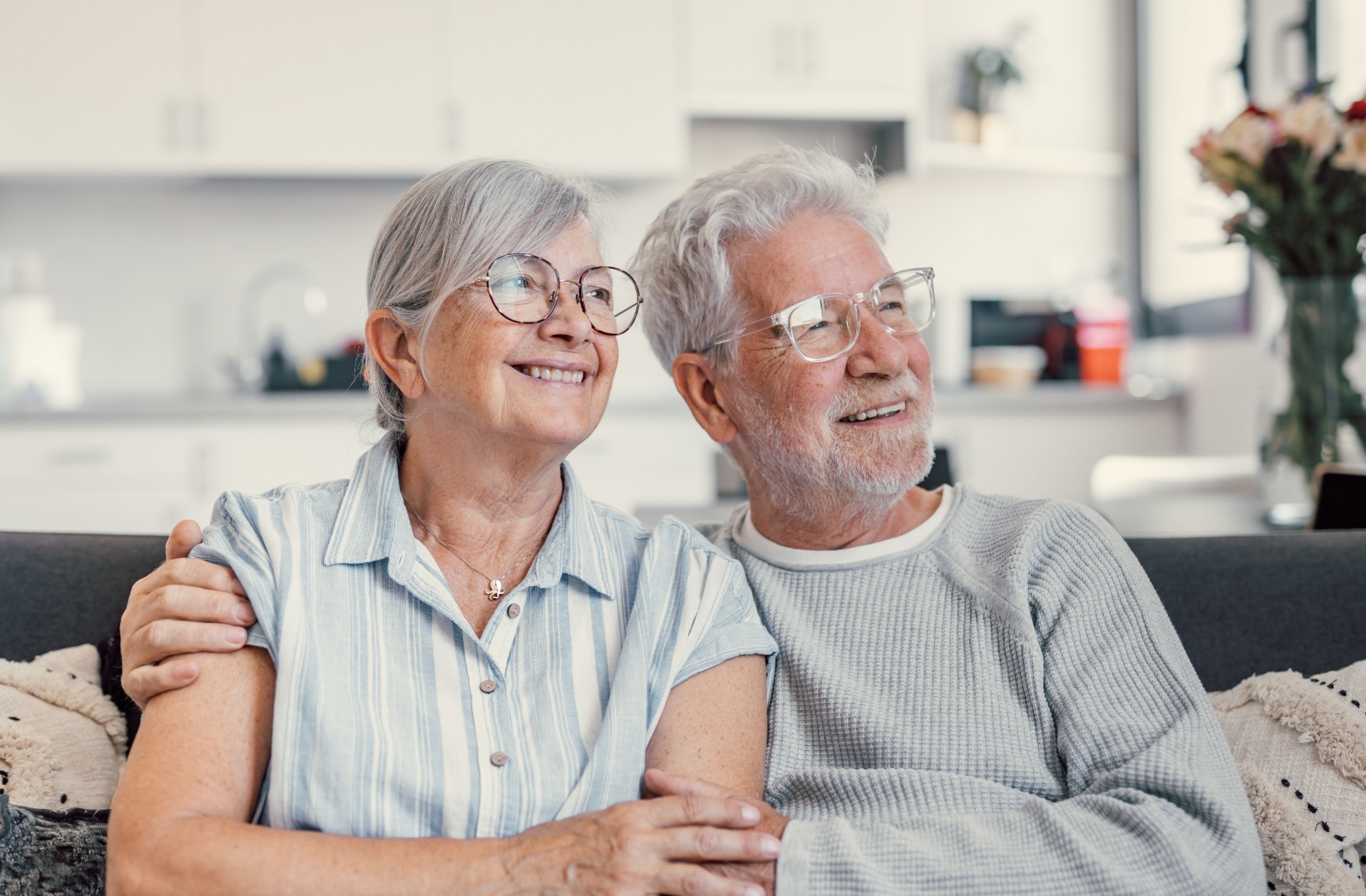 Two older adults sit on a couch with arms around one another and look out a window while smiling in their new senior living community