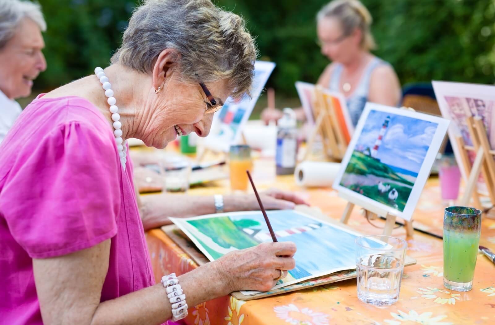 An older resident in assisted living paints a picture of a seaside scene while sitting outside with fellow residents
