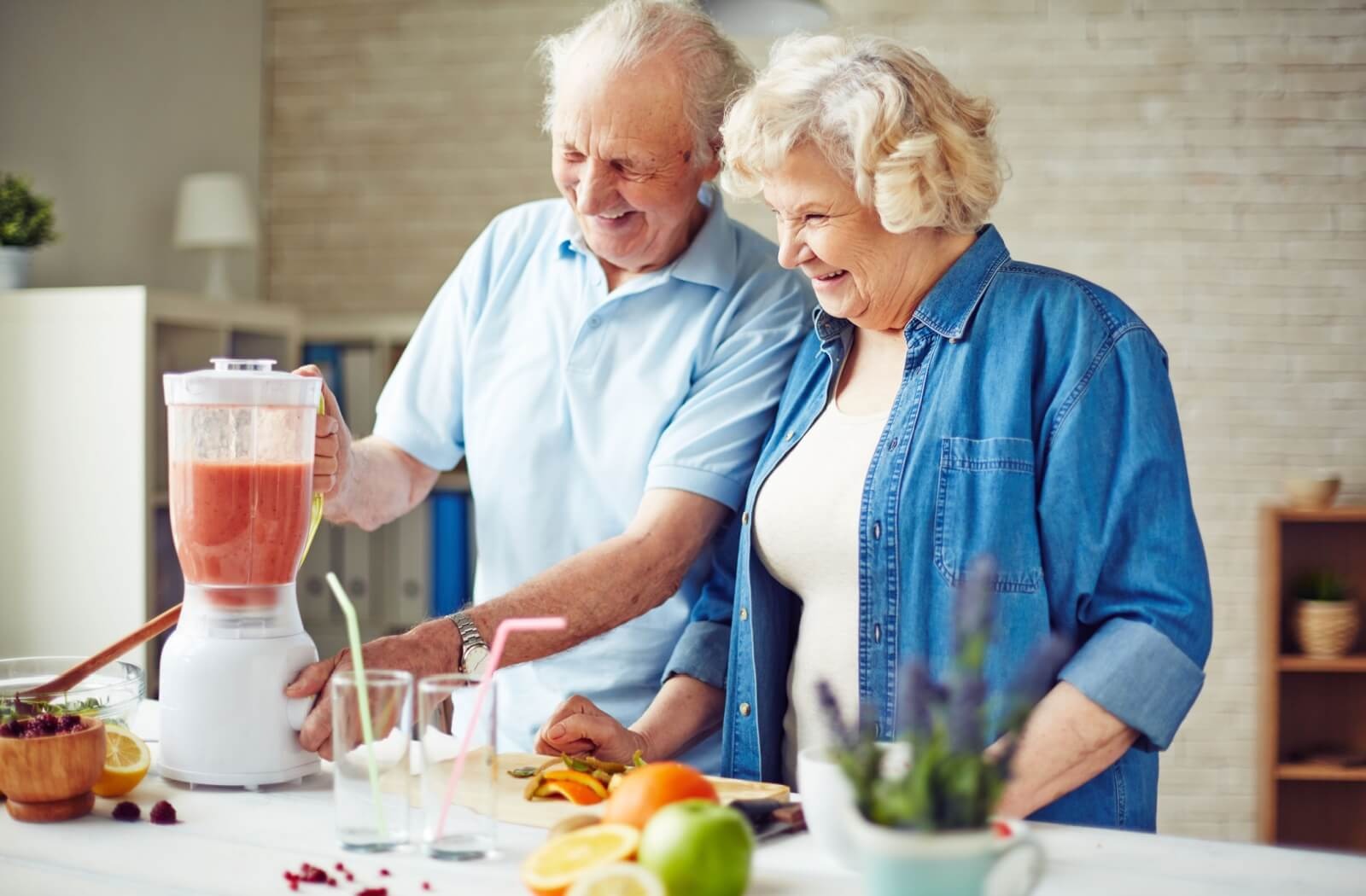 Two older adults smile while making a smoothie from fruit in their kitchen to help them stay hydrated
