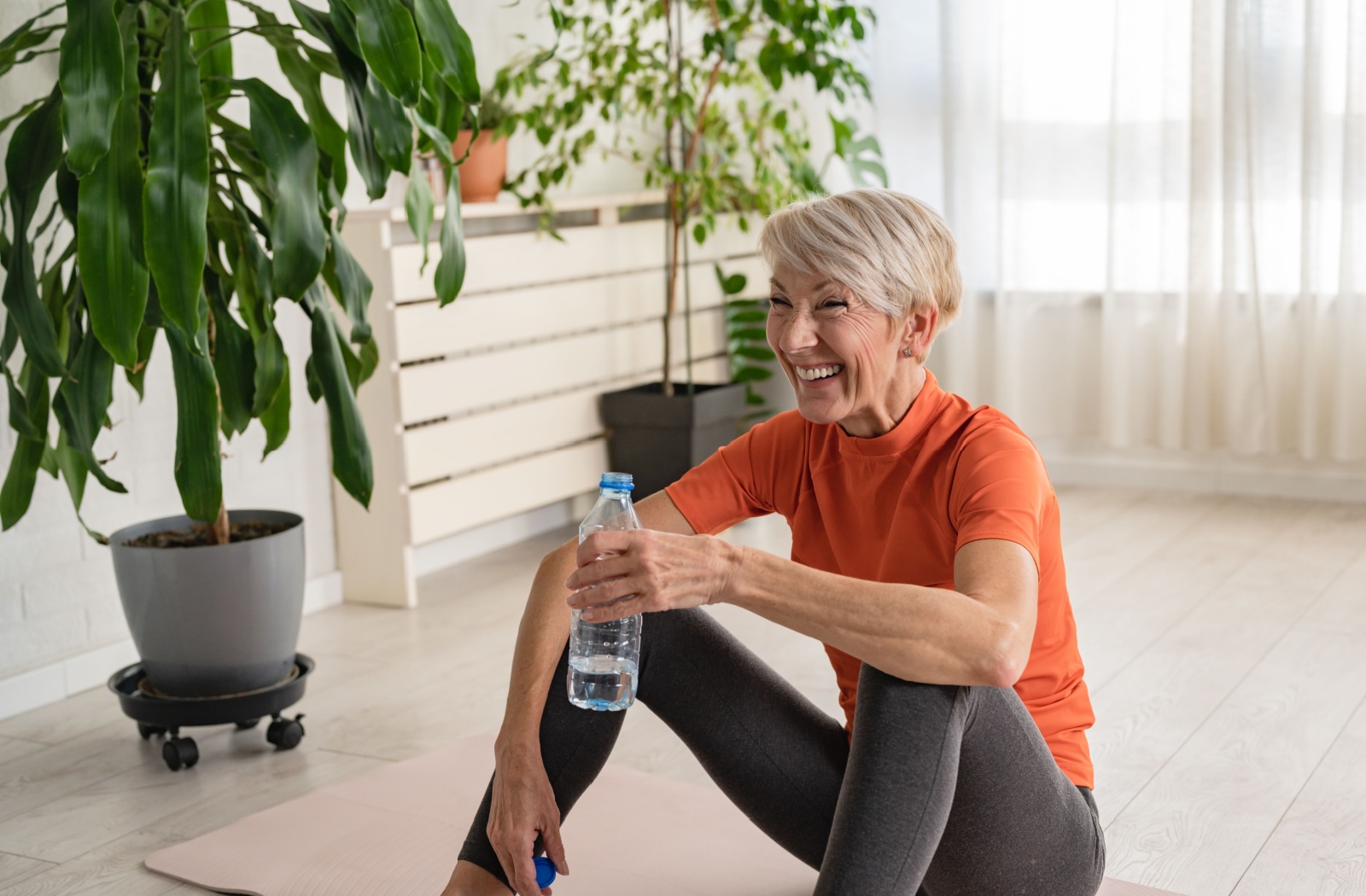 An older adult sits on the ground and sips a bottle of water while smiling during a break in a fitness class